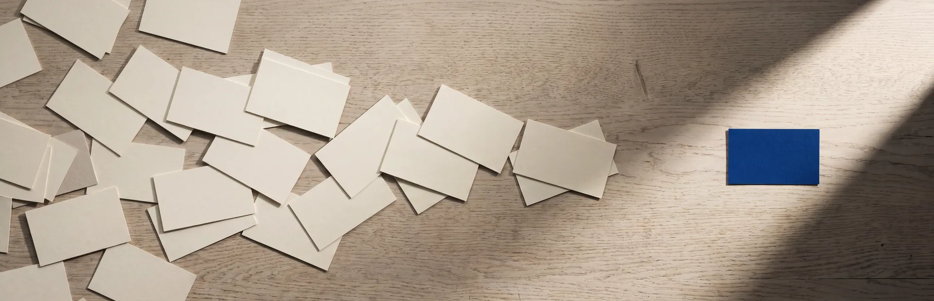 Dozens of blank cream cards scattered across a pale oak desk, with one deep blue card sitting alone in a shaft of sunlight